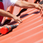 Workers on roof with electric drill installing red metal tile
